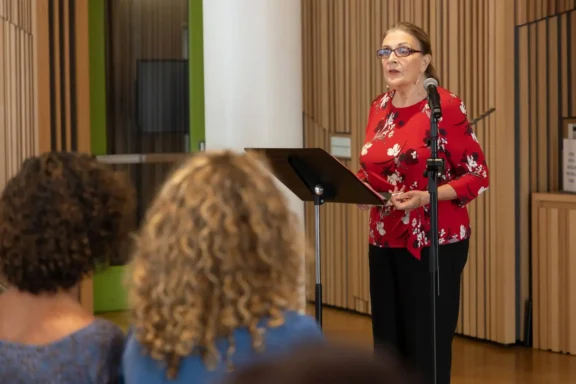 A woman in a red floral blouse speaks at a podium with a microphone in front of an audience in a modern indoor setting.