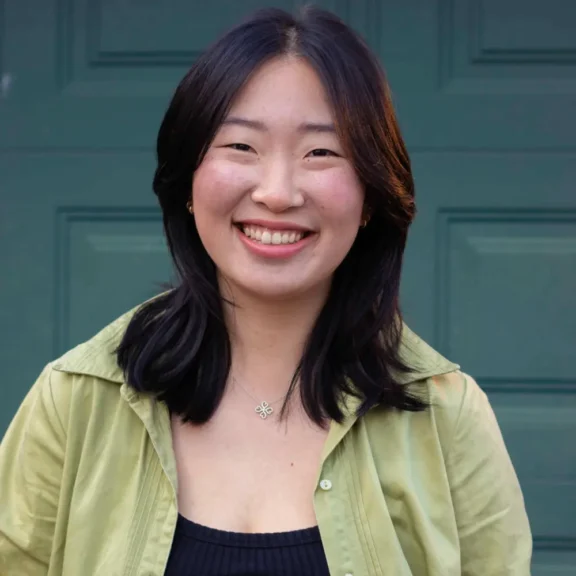 A woman with medium-length dark hair smiles at the camera, standing in front of a green garage door. She is wearing a light green shirt over a black top and a necklace.