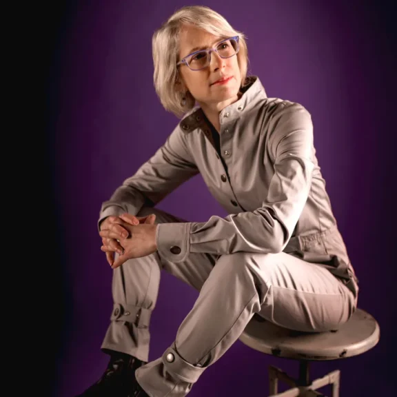 Person with short light hair and glasses sits on a stool, wearing a light gray jumpsuit, against a purple background.