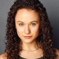 A woman with curly brown hair and blue eyes looks directly at the camera, wearing a sleeveless top and a thin necklace, against a plain gray background.
