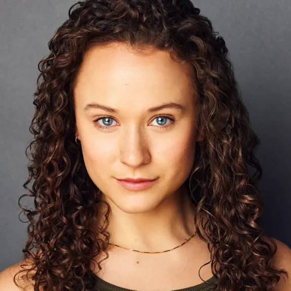 A woman with curly brown hair and blue eyes looks directly at the camera, wearing a sleeveless top and a thin necklace, against a plain gray background.