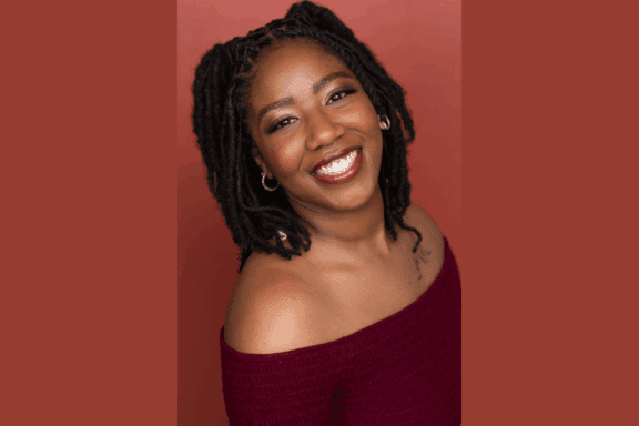 A woman with dark, shoulder-length locs smiles at the camera. She wears a burgundy off-shoulder top and small hoop earrings, with a reddish-brown background behind her.