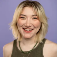 A person with short blonde hair, wearing a green sleeveless top and beaded necklace, smiles at the camera against a plain purple background.