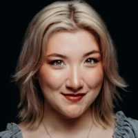 A person with short, layered blonde hair and light makeup smiles slightly at the camera against a dark background.