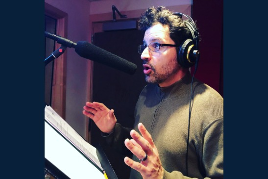 A man wearing headphones speaks into a studio microphone while reading from a script, gesturing with his hands.