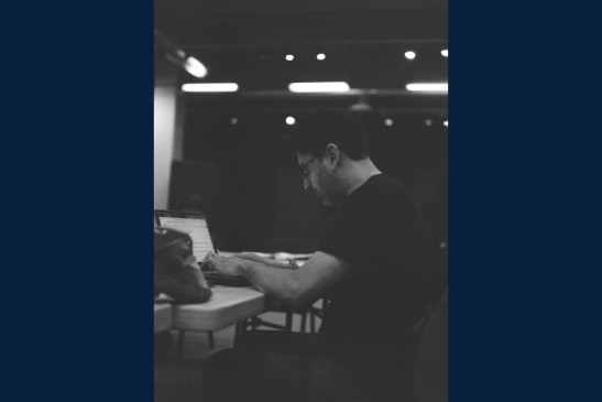 A person wearing glasses sits at a table, typing on a laptop in a dimly lit room.