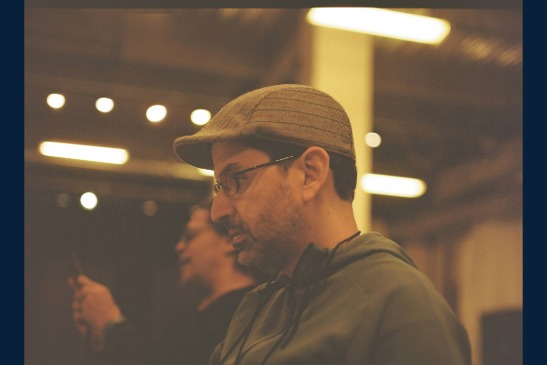 Two men indoors; the man in the foreground wears a flat cap and glasses, looking down, while the other man is blurred in the background holding a phone.