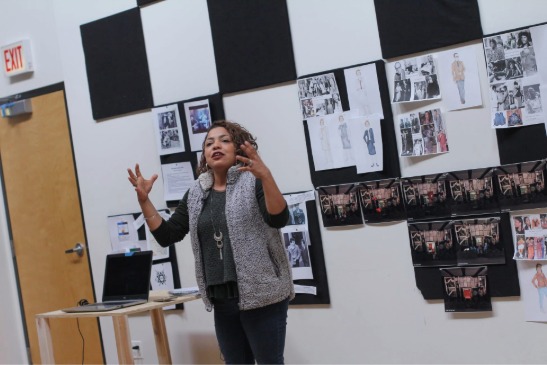 A person stands and gestures while presenting in front of a wall covered with fashion sketches and photographs, next to a laptop on a small table.
