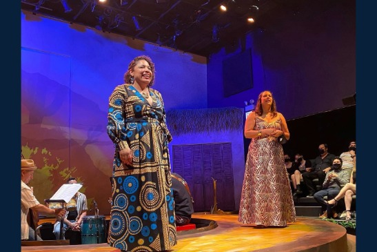 Two women stand and sing on a theater stage in colorful dresses; musicians and an audience are visible in the background.