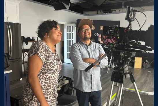 Two people stand next to a professional video camera on a tripod in a studio setting, looking at the monitor and smiling.