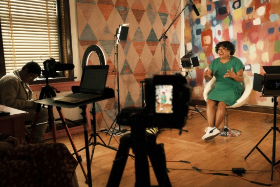 A woman in a green dress sits on a chair speaking in front of cameras and studio lights in a colorful room, recording a video interview.