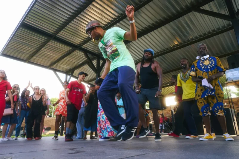 A group of people watches as a man in a green shirt and hat dances energetically under a pavilion, with others standing and clapping nearby.