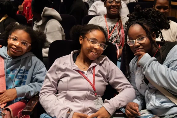 Three young people seated in an auditorium, all wearing glasses and casual clothing, smiling and looking at the camera. Other people are visible in the background.