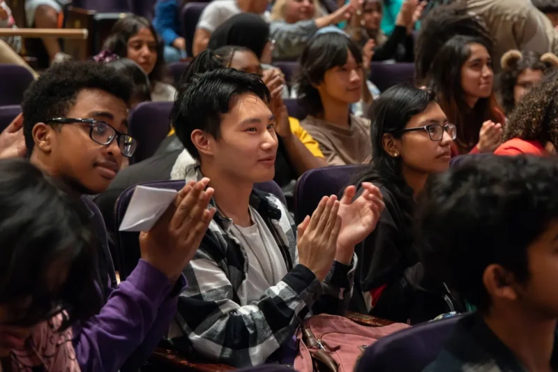 A group of people sitting in an auditorium, attentively watching and clapping during an event or presentation.