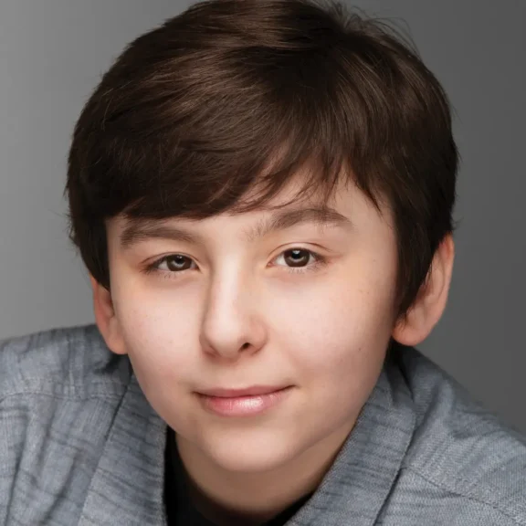 A young person with short brown hair and a neutral expression, wearing a gray sweater, poses against a plain gray background.