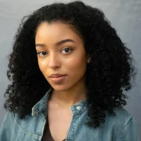 A woman with curly black hair wearing a denim jacket looks directly at the camera with a neutral expression against a plain background.