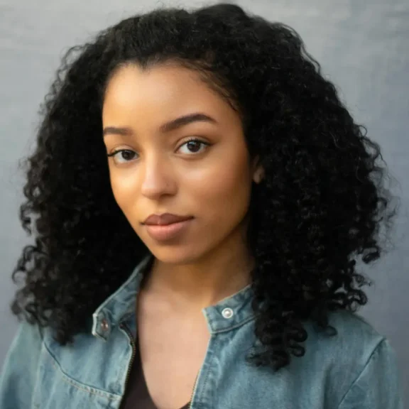 A woman with curly black hair wearing a denim jacket looks directly at the camera with a neutral expression against a plain background.
