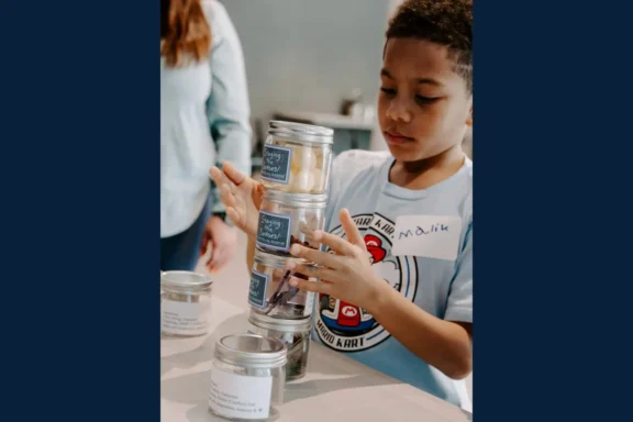 A young boy stacks labeled glass jars on a table while a woman stands nearby in the background.