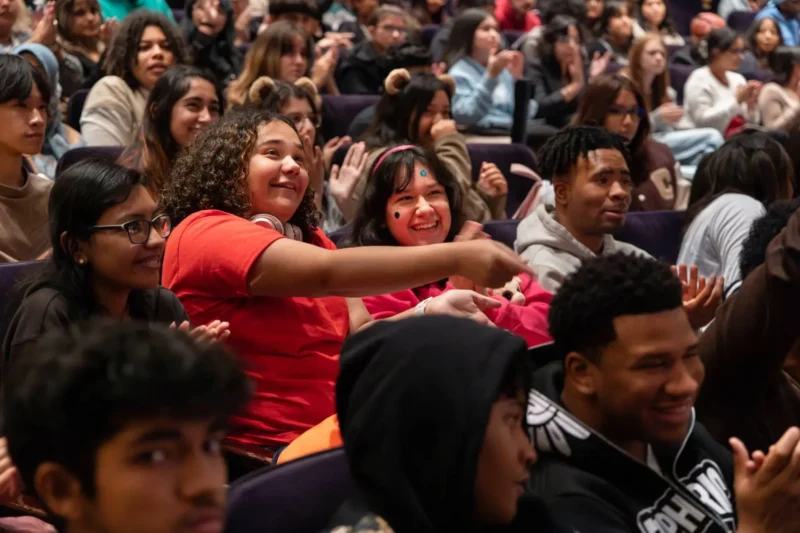 A large group of diverse students sit in an auditorium, some smiling, pointing, and clapping, while others watch attentively.