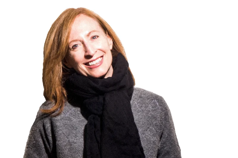 A woman with red hair, wearing a gray sweater and a large black scarf, smiles while facing the camera against a white background.
