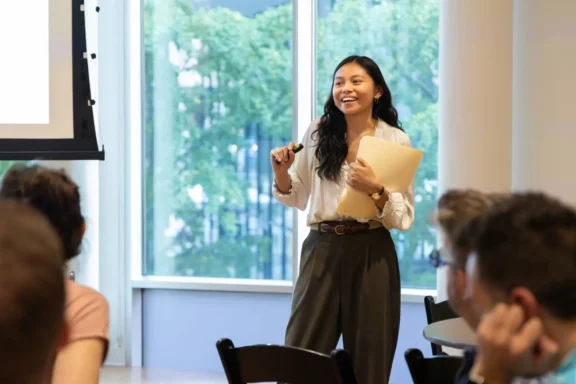 A woman is standing and speaking in front of a group, holding papers and a marker, with large windows and trees visible in the background.