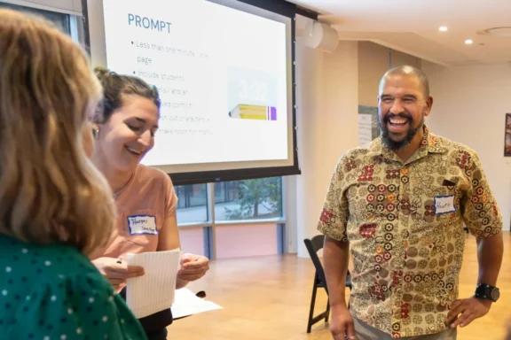 Three people interact and smile during a workshop; a presentation slide is displayed in the background.