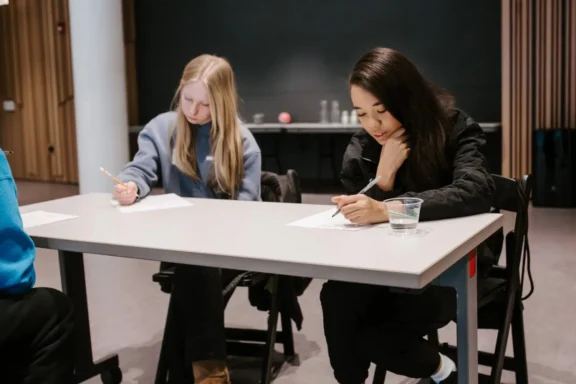 Two young women sit at a table writing on paper with pencils. A glass of water is on the table. The background features wooden paneling and a dark wall.
