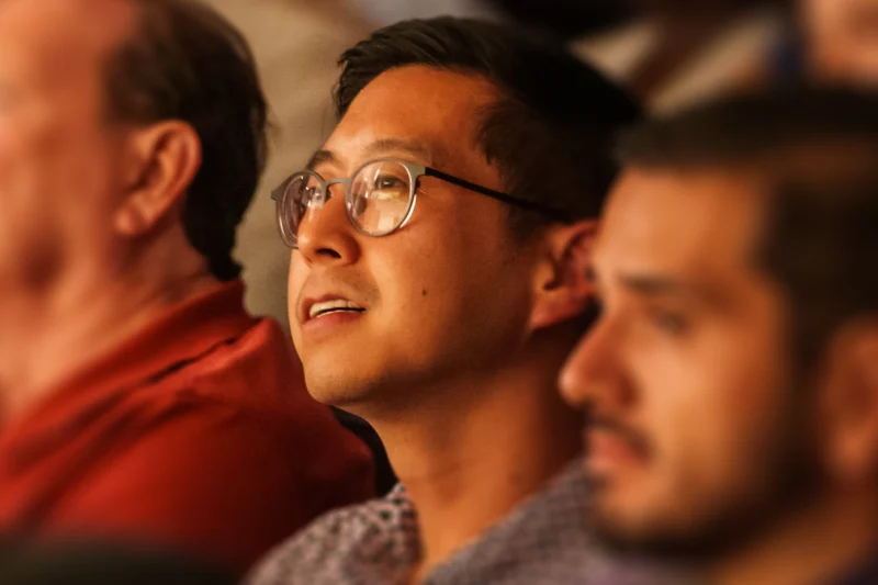 A man with glasses sits among others, looking forward with a slight smile. The image focuses sharply on him while the surroundings are blurred.