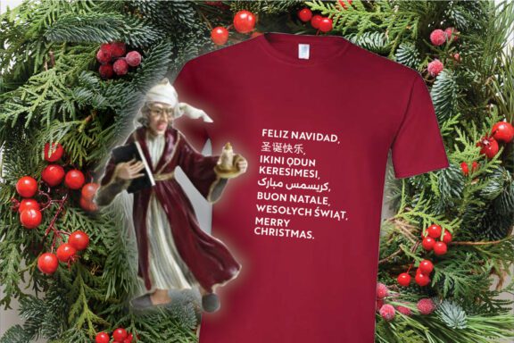A maroon T-shirt with "Merry Christmas" in multiple languages is displayed beside a figurine of an elderly person holding a lantern, against a festive background of greenery and red berries.