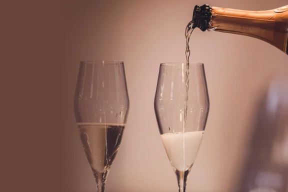 A close-up of champagne being poured from a bottle into one of two clear wine glasses, with a plain background.