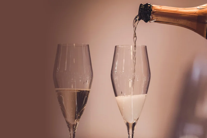A close-up of champagne being poured from a bottle into one of two clear wine glasses, with a plain background.