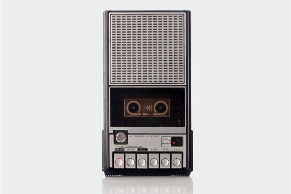 Front view of a vintage portable cassette tape recorder with buttons, speaker grille, and cassette slot visible against a neutral background.