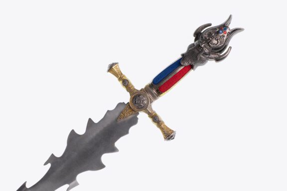 A decorative sword with a wavy blade, ornate gold crossguard, and a handle featuring red and blue inlays and an intricate sculpted face.