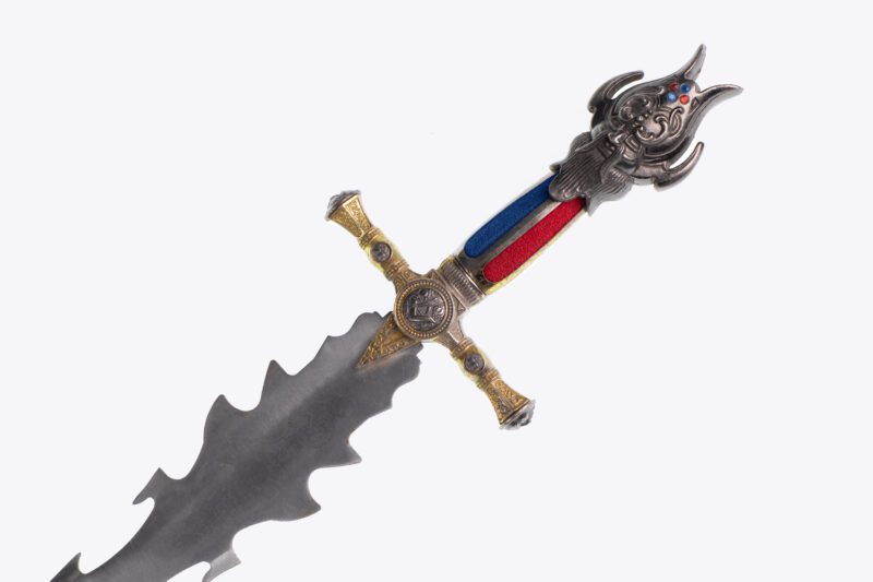 A decorative sword with a wavy blade, ornate gold crossguard, and a handle featuring red and blue inlays and an intricate sculpted face.