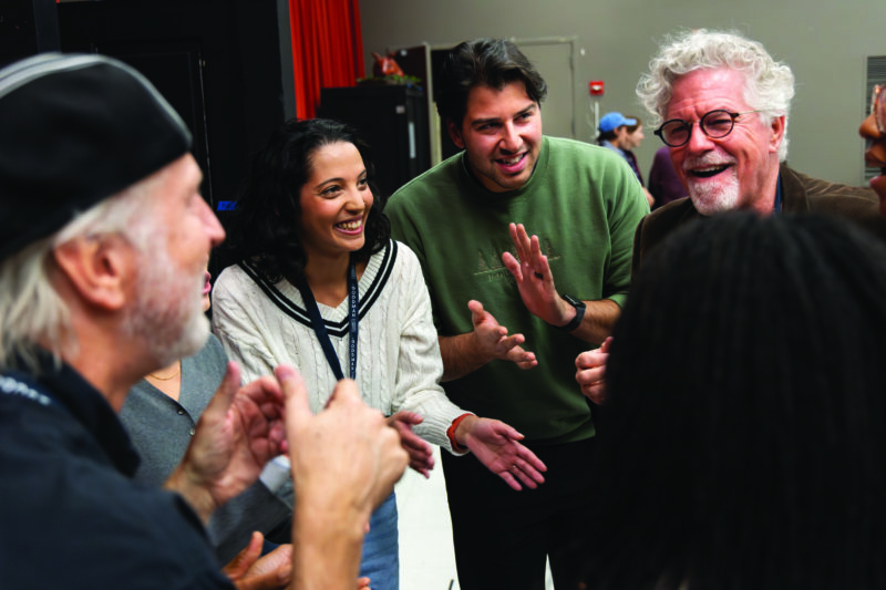 A group of five adults stand in a circle indoors, smiling and clapping, appearing to enjoy a lively conversation or activity.