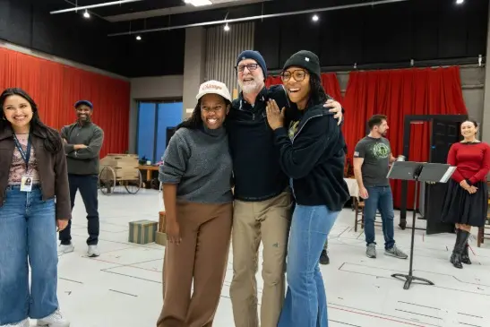 A group of people stand and pose together in a rehearsal space, smiling and interacting, with music stands and props in the background.