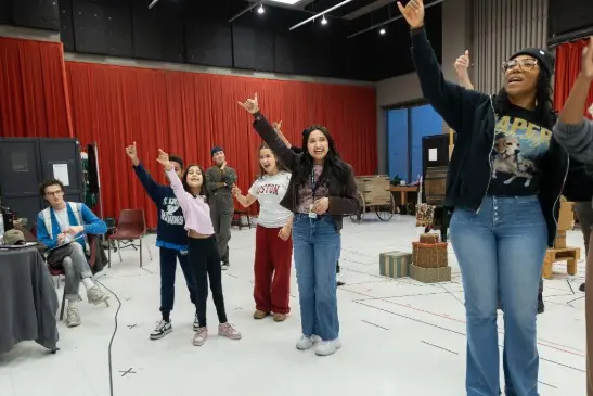 A group of people stand in a rehearsal space, smiling and raising one arm enthusiastically while others sit and watch in the background.