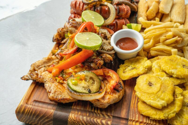 A wooden board with grilled chicken topped with vegetables and lime, fried plantains, French fries, and a small cup of dipping sauce.