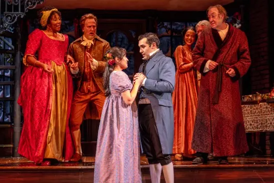 A group of actors in period costumes perform on stage, with two people in the center holding hands while others react expressively in the background.