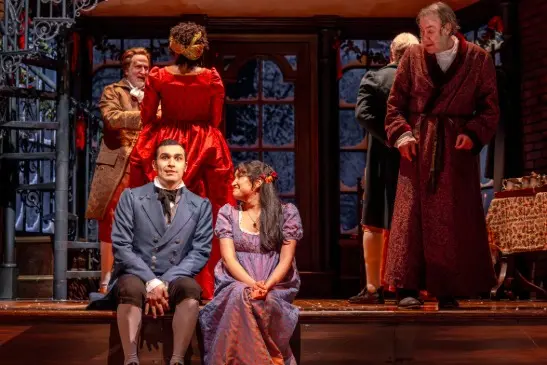 Six actors in period costumes perform on stage, with two sitting in the foreground and four standing in the background against a winter-themed set.