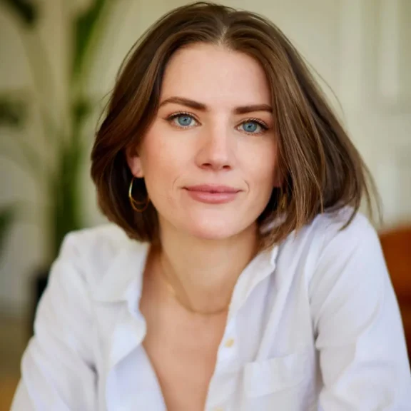 A woman with short brown hair and blue eyes wearing a white shirt and gold hoop earrings, looking directly at the camera with a neutral expression.