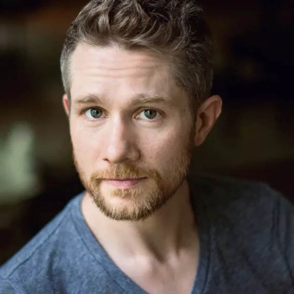 A man with short light brown hair and a trimmed beard, wearing a grey V-neck shirt, looks directly at the camera against a dark background.