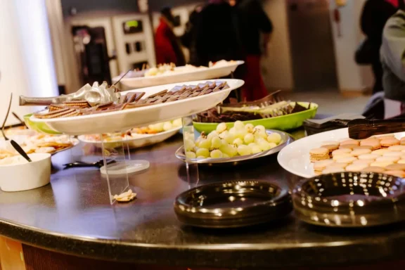 Plates of assorted desserts, including cookies, macarons, chocolate, and green grapes, are arranged on a round buffet table.