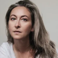 A person with long, wavy, gray hair and light skin looks directly at the camera with a neutral expression against a plain background.