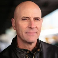 A middle-aged man with a shaved head wearing a black leather jacket looks at the camera with a neutral expression.