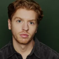 A young man with short, wavy red hair and a trimmed beard poses against a dark green background, wearing a dark corduroy shirt and looking directly at the camera.