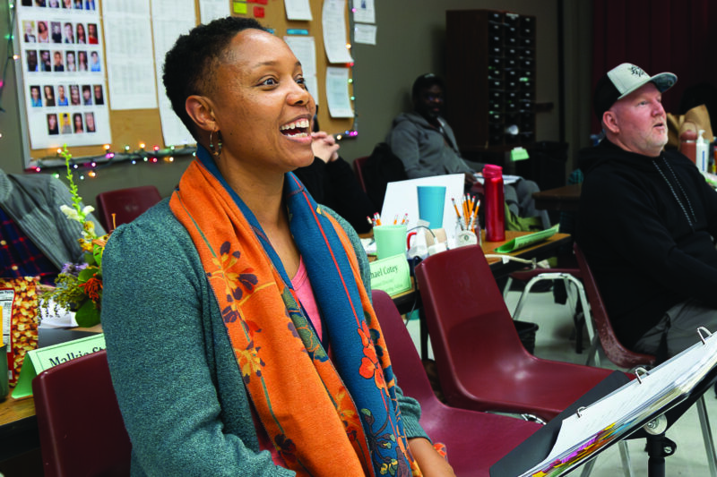 A woman sits smiling in a classroom setting with papers and nameplates on desks; other people are seated nearby, and a bulletin board is visible in the background.