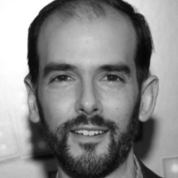 Black and white close-up of a man with short dark hair, a beard, and a slight smile, facing the camera against a blurred background.
