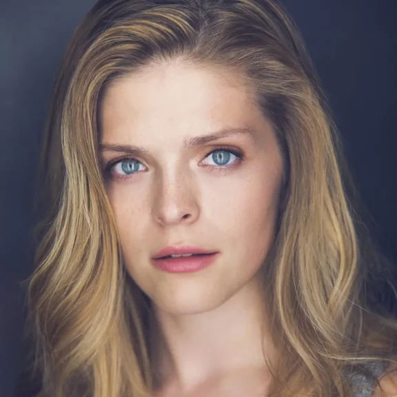 Woman with long blonde hair and blue eyes looking directly at the camera, neutral expression, dark background.