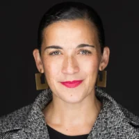 A woman with dark hair, wearing geometric earrings and a patterned jacket, faces the camera against a black background.